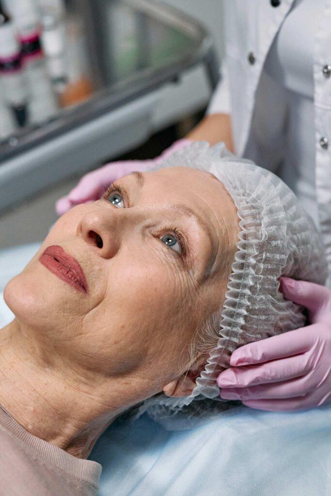 Dr. Maeve O'Doherty, an older woman with a hair cap, receiving a facial examination or treatment from a healthcare professional wearing pink gloves in a clinical setting.